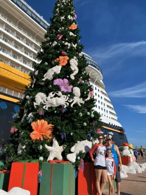 family of three at the base of large Christmas tree with cruise ship behind them and the tree