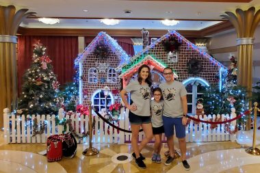 family of three in matching grey shirts with lit up gingerbread house behind them