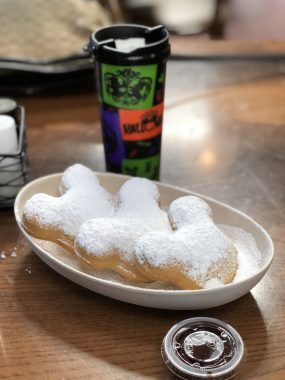 three mickey shaped beignets with coffee in green, orange and purple cup behind