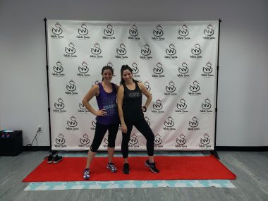 Two smiling women pose in front of a photo backdrop at a dance studio