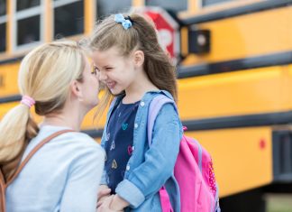 Mom gives daughter eskimo kiss before the girl boards school bus. The girl is excited about her first day of kindergarten. The school bus is in the background.