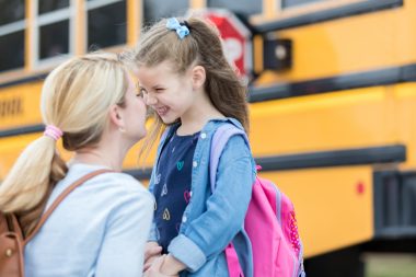 Mom gives daughter eskimo kiss before the girl boards school bus. The girl is excited about her first day of kindergarten. The school bus is in the background.