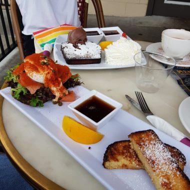 two plates of food on table with a cup of coffee. one plate is rectangular and has poaches egg on top of bed of lettuce and bread with parika sprinkled on top, syrup in a square bowl and orange slice next to it, then french toast cut into strips for dipping. The other plate has french toast with chocolate icecream and dollop of whipped cream to the side. 