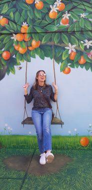 woman is laughing as she leans against a mural of an orange tree with a swing. Woman is pretending to sit on the painted swing