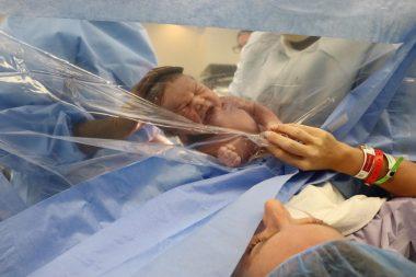 Mom just gave birth via c-section, baby is behind clear plastic as mom reaches out to hold baby's hand. She is looking at him. 