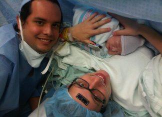 mom just having given birth via c-section to a baby boy. Mom is wearing a medical cap, with a black temperature sticker on her forehead, black glasses and holding baby. Dad is off to the left. Both are smiling.