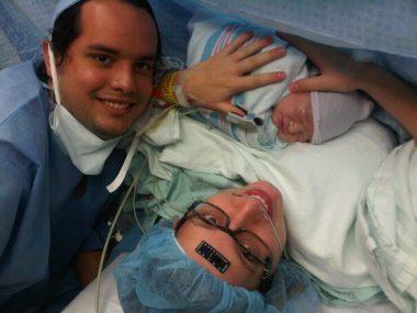 mom just having given birth via c-section to a baby boy. Mom is wearing a medical cap, with a black temperature sticker on her forehead, black glasses and holding baby. Dad is off to the left. Both are smiling. 