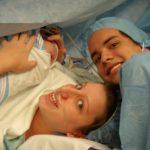 mom just gave birth via c-section. mom is toward the bottom of the screen smiling faintly, baby is wrapped up in a blanket, moms hands are over his body, dad is toward the right of the photo smiling.