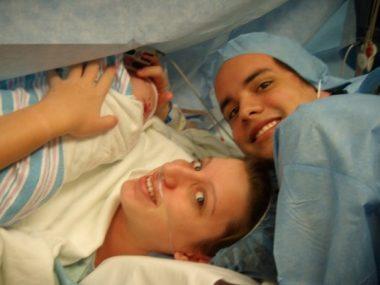 mom just gave birth via c-section. mom is toward the bottom of the screen smiling faintly, baby is wrapped up in a blanket, moms hands are over his body, dad is toward the right of the photo smiling. 