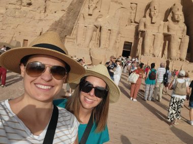 two women wearing sunglasses on a sunny day smiling at the camera, with a close up of one of the pyramids of egypt behind them. 