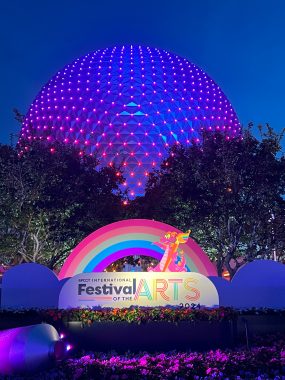 Festival of the Arts sign with Figment and rainbow in from of blue and purple lit up Spaceship Earth.