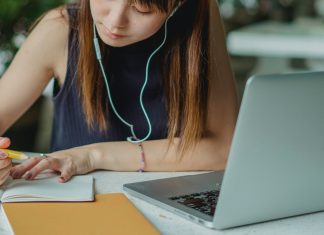 girl writing in notebook with earbuds and a computer in front of her, going through college asmission