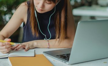 girl writing in notebook with earbuds and a computer in front of her, going through college asmission