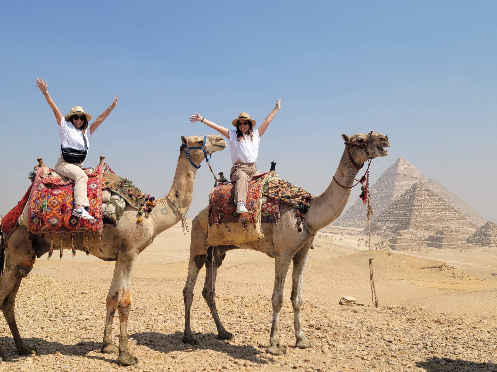 two women on top of two camels with the pyramids of Egypt behind them
