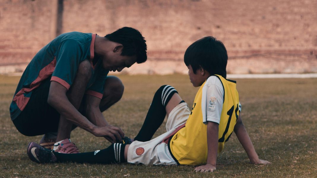 young athletes sitting on ground, while older athlete or coach teaches him about sports safety and ties his shoes